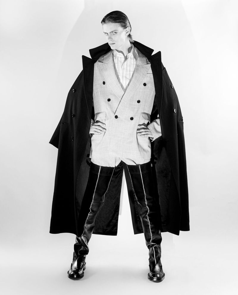 Black and white studio shot of androgynous fashion model with black overcoat, double-breasted jacket, high boots, hands on hips, gazing at viewer.