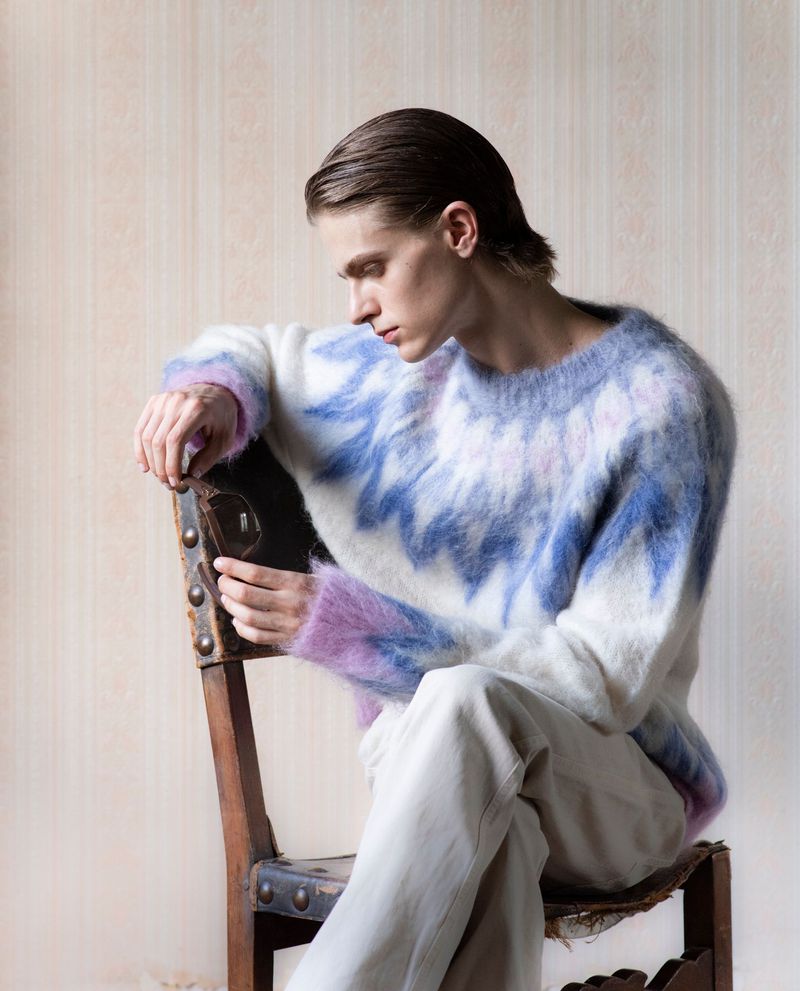 A fair-skinned young man sits on a vintage chair in a studio, wearing a fuzzy blue-and-white sweater and light pants in soft, diffused light.