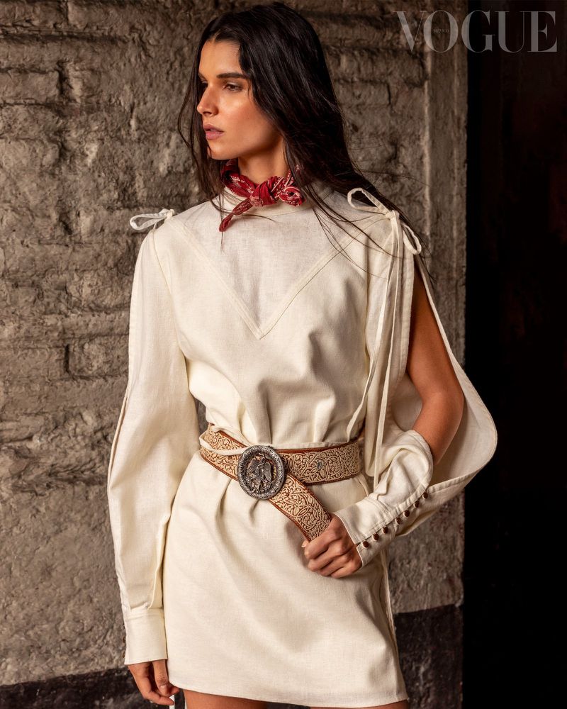 Model wearing an off-white split-sleeve mini dress with a carved leather belt and red neck scarf against a stone wall.