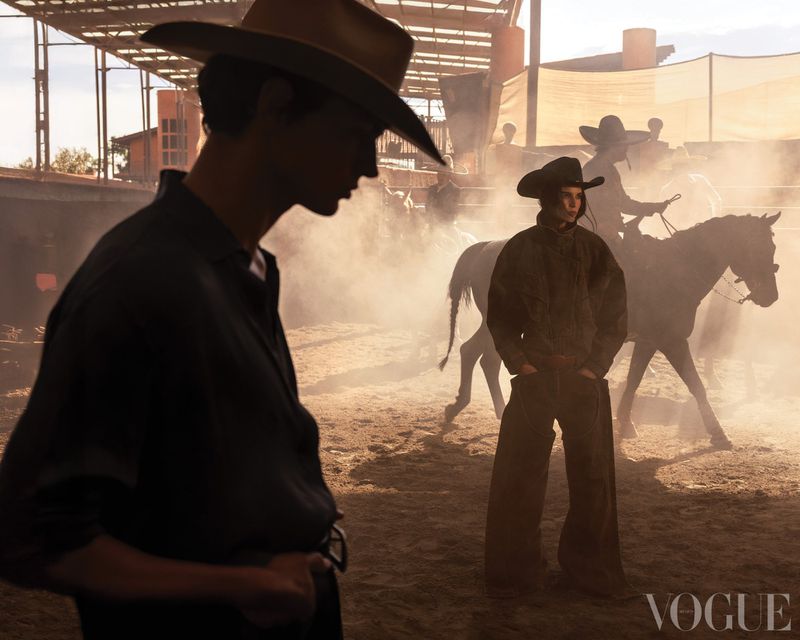 Cowboys on horseback in a dusty outdoor arena, illuminated by sunlight. Two men in foreground in Western wear. Vogue logo at bottom.