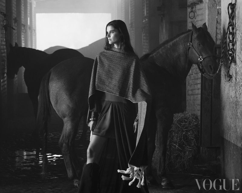 Monochrome shot of a woman in a poncho and skirt posing with two horses in a stable, soft lighting, and a Vogue logo at the bottom.