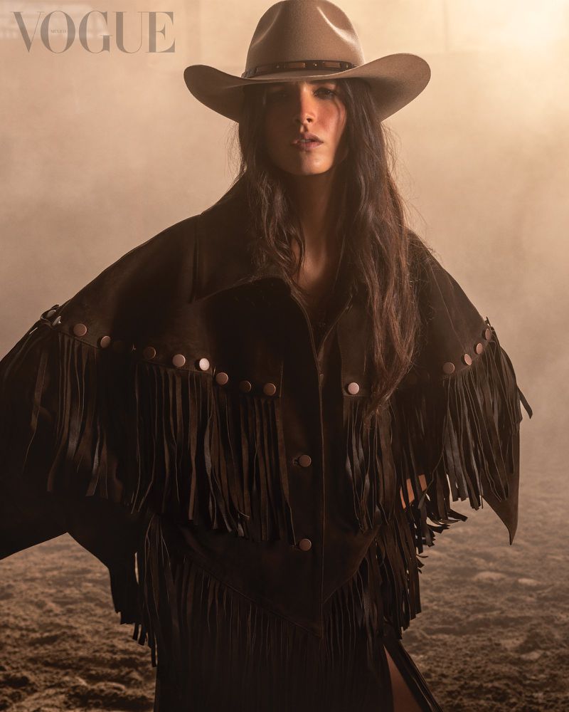 Brunette woman in a tan cowboy hat and fringed jacket poses for a Vogue fashion editorial with warm studio lighting and a textured background.