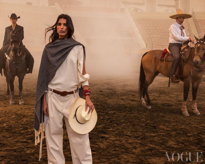 Editorial fashion shot featuring a woman in white holding a hat, posed in a bullring with two other people on horseback in the background.