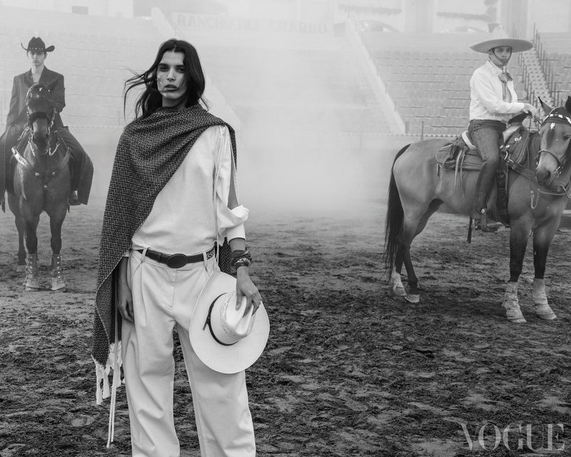 Black and white outdoor fashion shot of a woman in white clothing holding a hat, with two men riding horses in the background.