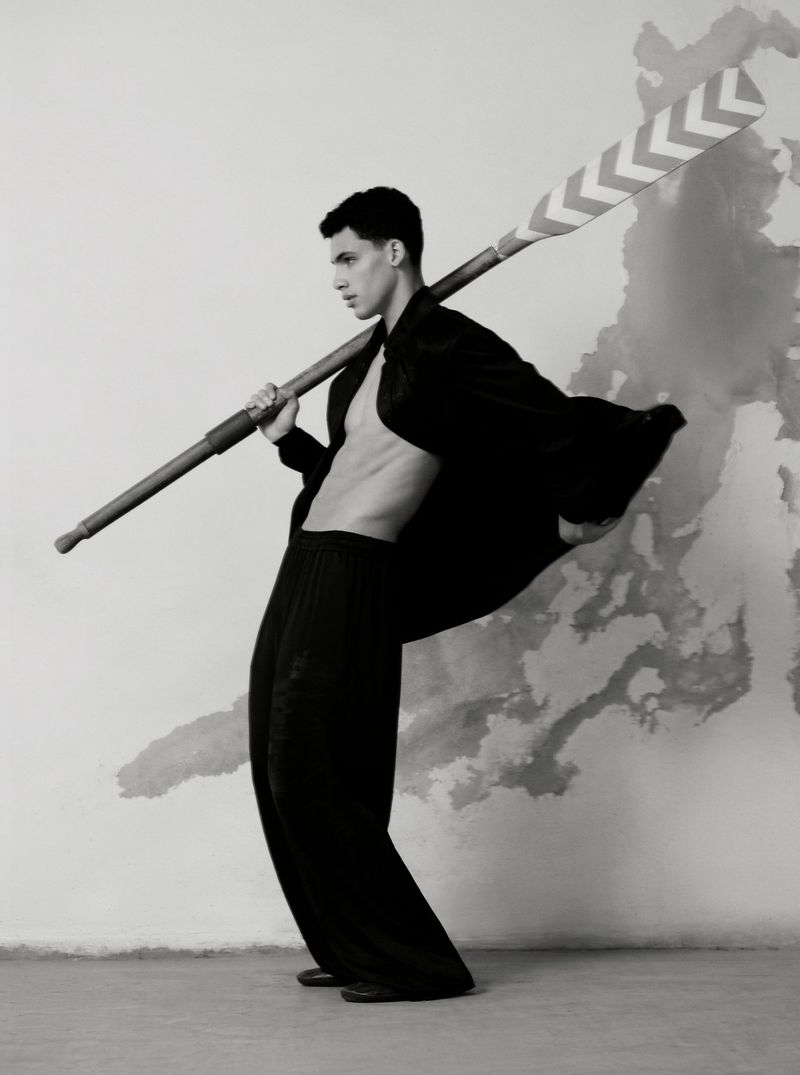 Young man in an open black blazer, holding an oar on his shoulder, poses in front of a textured wall in a studio setting.