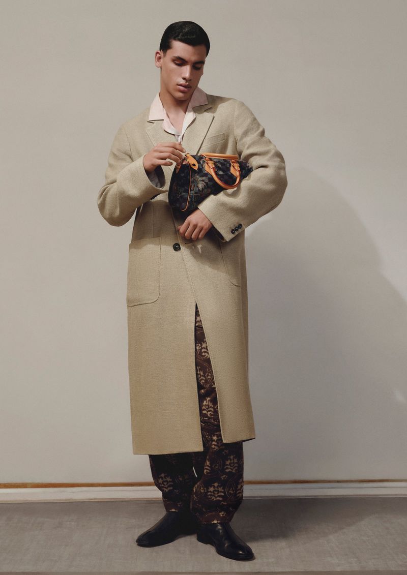 An adult man posing indoors in a full-length shot, wearing a long beige coat, patterned pants, and black shoes, and holding a black bag.
