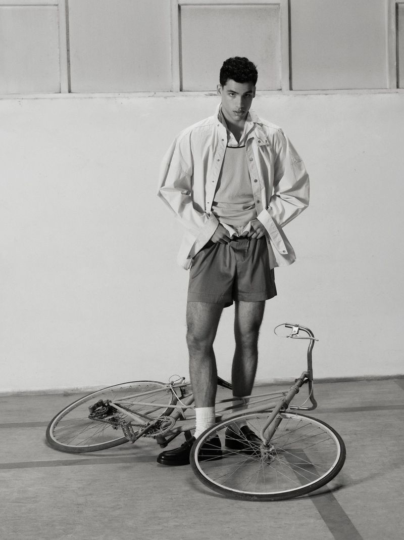 Monochrome portrait of a male model in shorts and a jacket standing next to a bicycle in a studio with a light-grey backdrop.