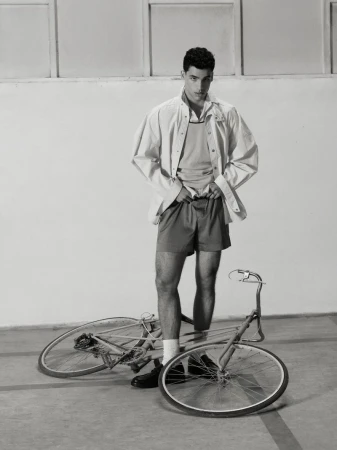 Monochrome portrait of a male model in shorts and a jacket standing next to a bicycle in a studio with a light-grey backdrop.