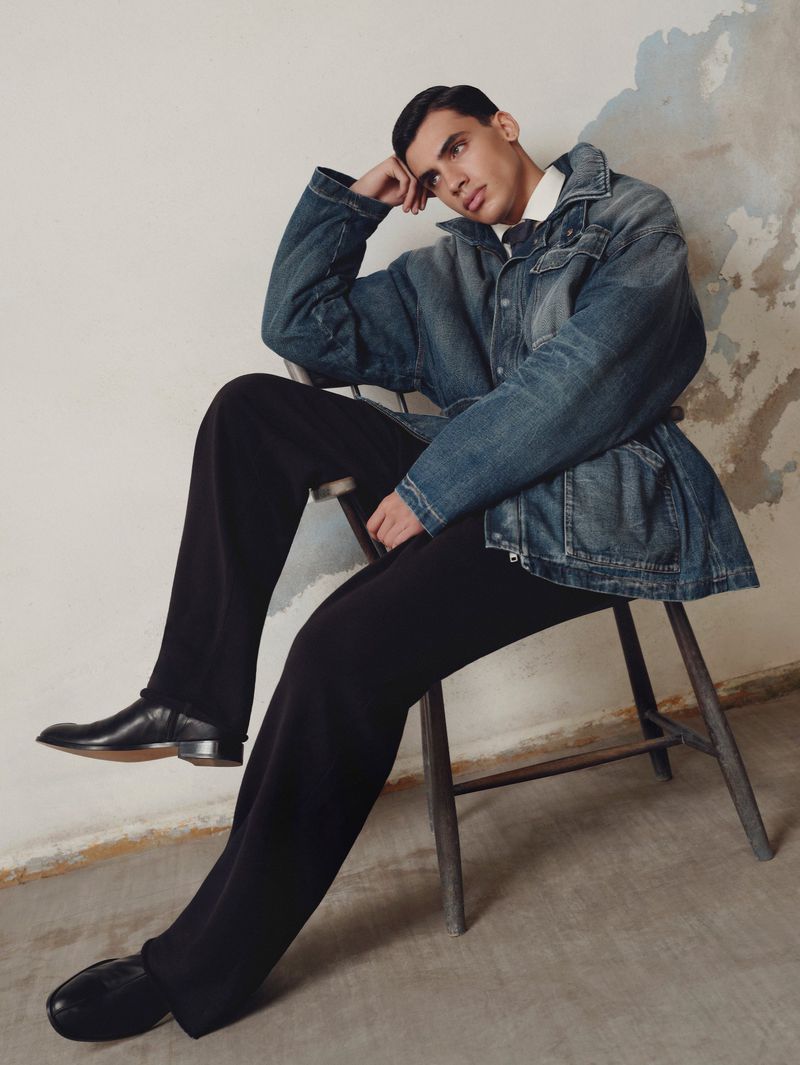 Stylish man sitting on a wooden chair in studio. He's wearing a denim jacket, dress shirt, black pants, and black leather shoes.