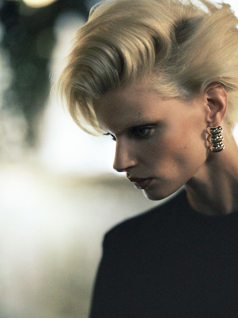 Close-up studio portrait of an adult woman with blonde hair wearing black top and gold earrings with a blurred gradient background.