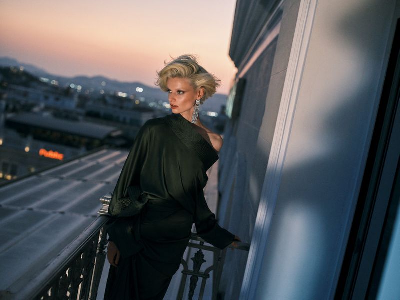 Adult woman poses in an elegant green dress on a balcony overlooking a cityscape at dusk. She wears large chandelier earrings.