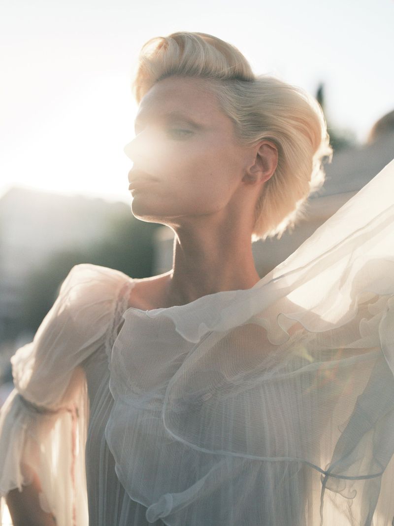Blond woman in a white dress poses outdoors in bright sunlight, creating a bright, ethereal aesthetic with buildings in the background.