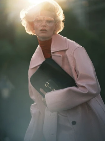 A fair-skinned woman with blonde hair, wearing aviator glasses and a pink coat, poses in front of a blurred green backdrop with bright sun flares.