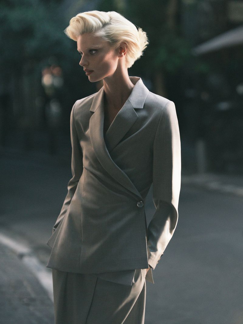 Fashion portrait of a woman with blonde hair wearing a light grey suit in an urban environment with soft, diffused lighting.