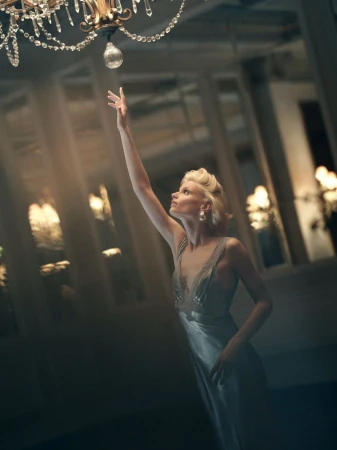 Woman in a silk slip dress reaches for a crystal chandelier. Soft lighting creates a dramatic atmosphere. Beauty and fashion are prominent.