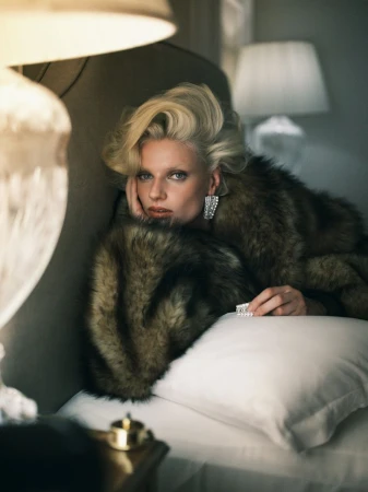 Model Mila van Eeten wearing a voluminous brown fur coat and sparkling crystal earrings while lying on a bed with a white pillow.