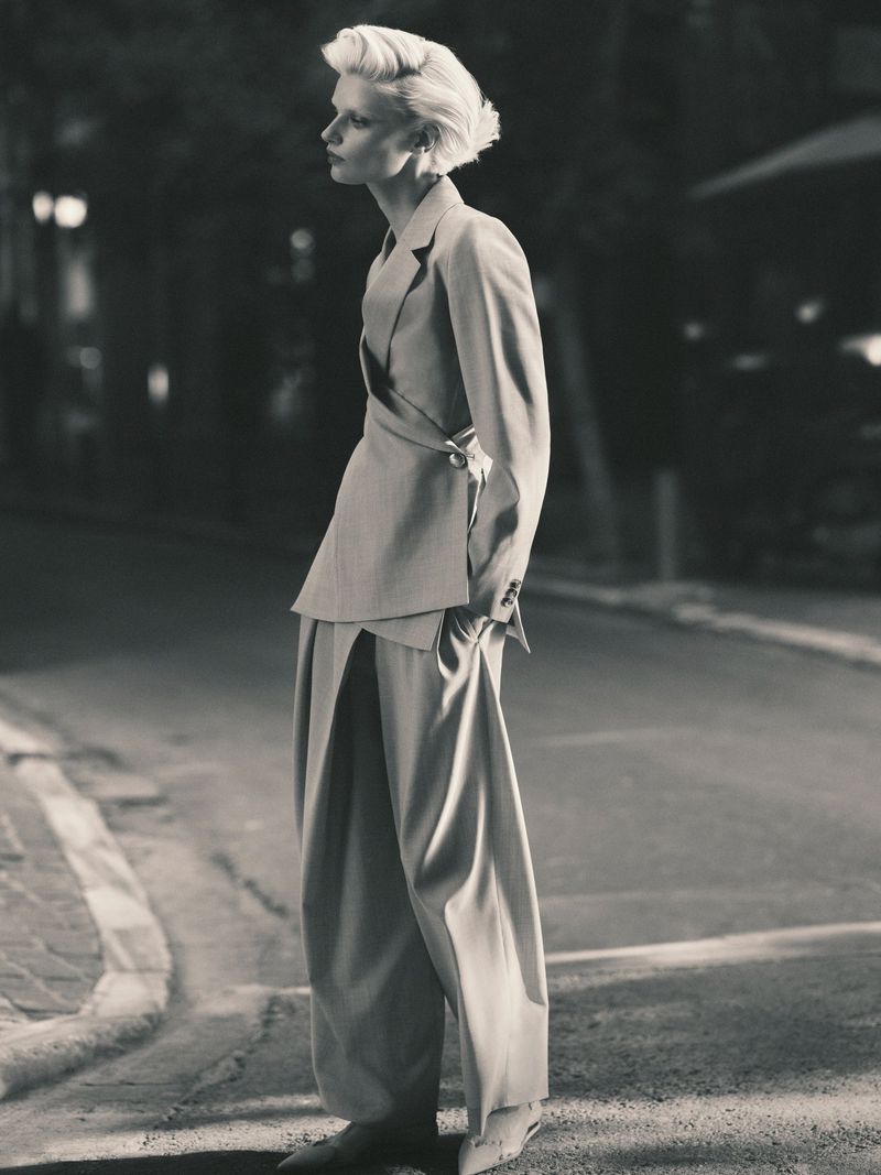 A woman poses in a gray suit with wide-legged pants on a city street, showcasing an editorial fashion look with blurred background lights.