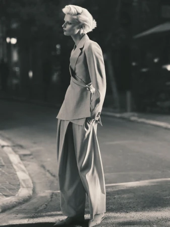 A woman poses in a gray suit with wide-legged pants on a city street, showcasing an editorial fashion look with blurred background lights.