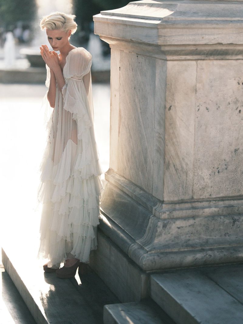 Elegant woman in flowing white gown stands near marble structure outdoors, captured in soft, natural light with fountains visible in background.