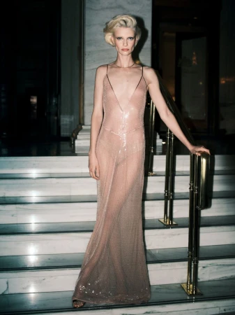 Model Mila van Eeten wearing a sheer shimmering blush evening dress while standing on a marble staircase holding a brass handrail.