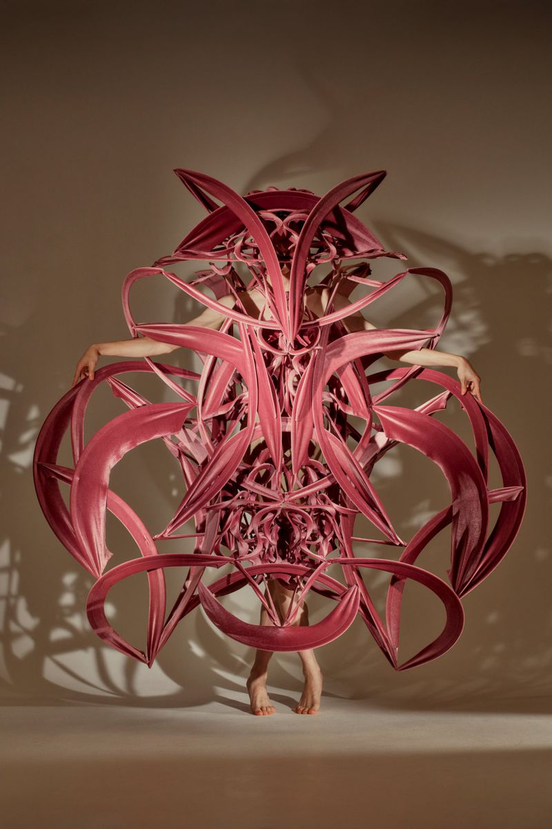 Adult female posing in an abstract, sculptural pink dress against a neutral backdrop in a studio setting with creative fashion.