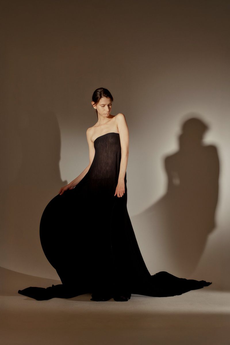A woman poses in a flowing black dress against a neutral background, with soft lighting enhancing the fabric's texture and the model's silhouette.