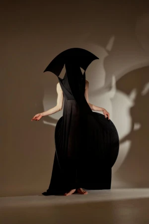 Fashion photography showcases a woman in a flowing black dress with architectural shapes, set against a light-colored studio backdrop.