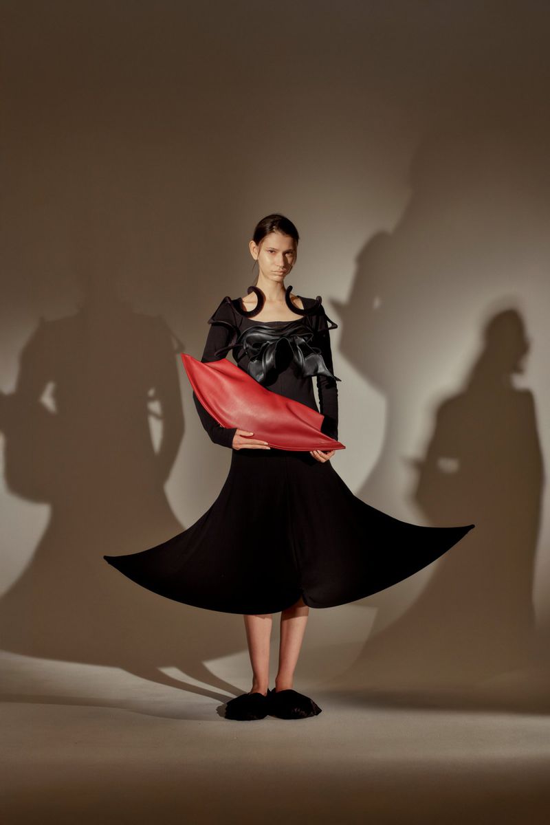 Fashion model stands in a studio wearing a black dress and holding a red bag, creating a dramatic pose with shadows in the background.