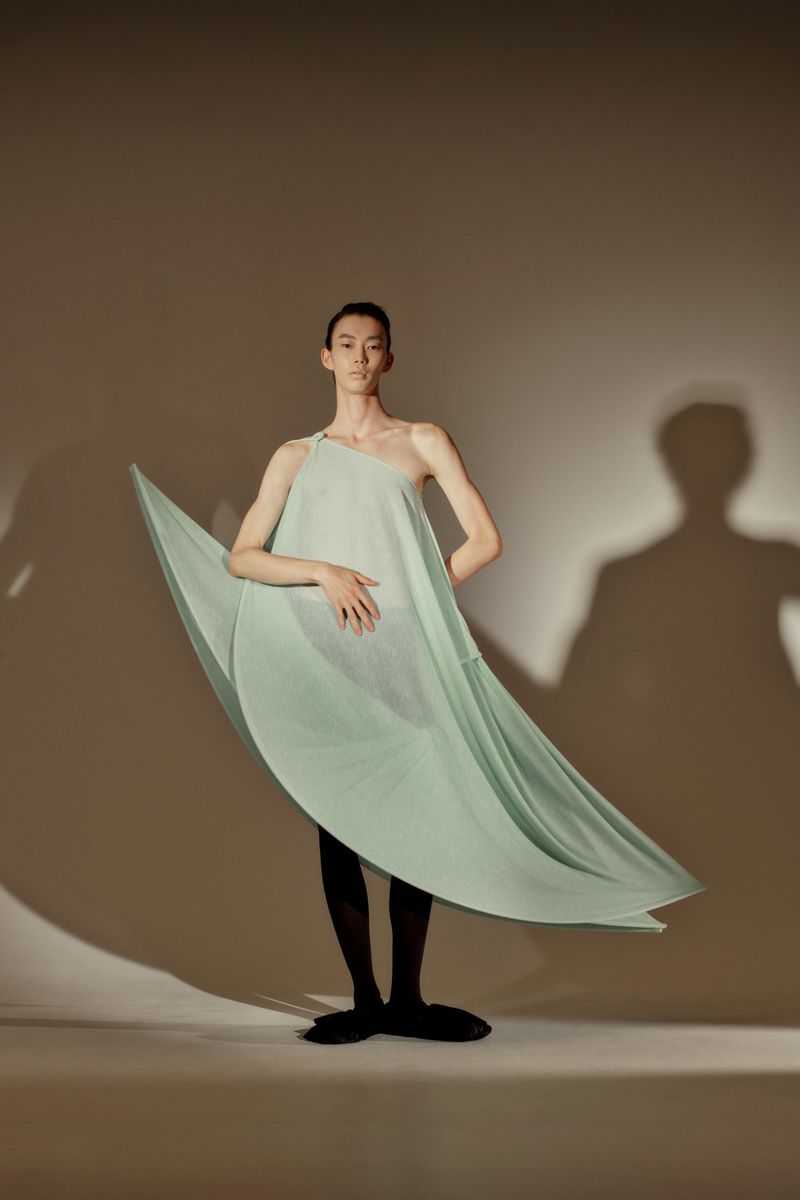 Adult male in studio, wearing a light green one-shoulder dress and black tights, standing in front of a beige background with shadows.