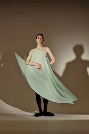 Adult male in studio, wearing a light green one-shoulder dress and black tights, standing in front of a beige background with shadows.