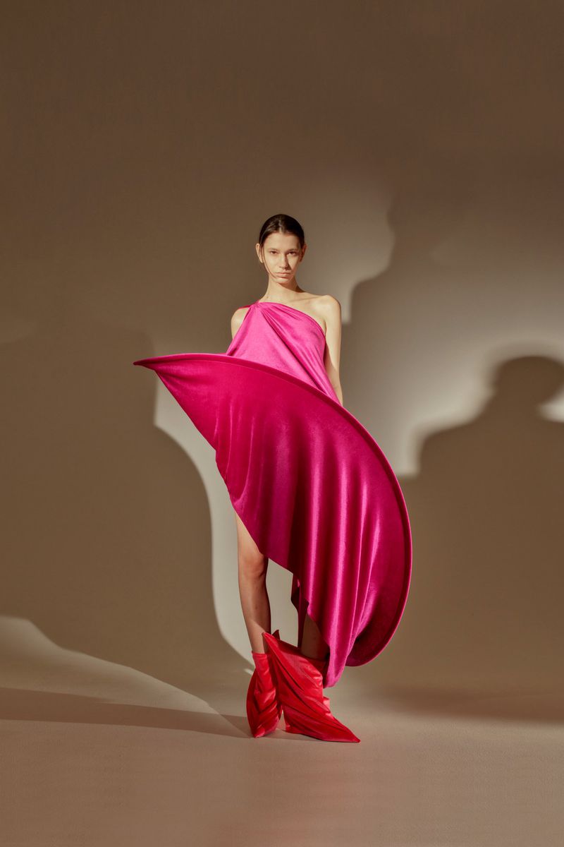 A woman poses in a pink one-shoulder dress with flowing fabric, paired with red feather-like shoes, in a fashion studio shoot.