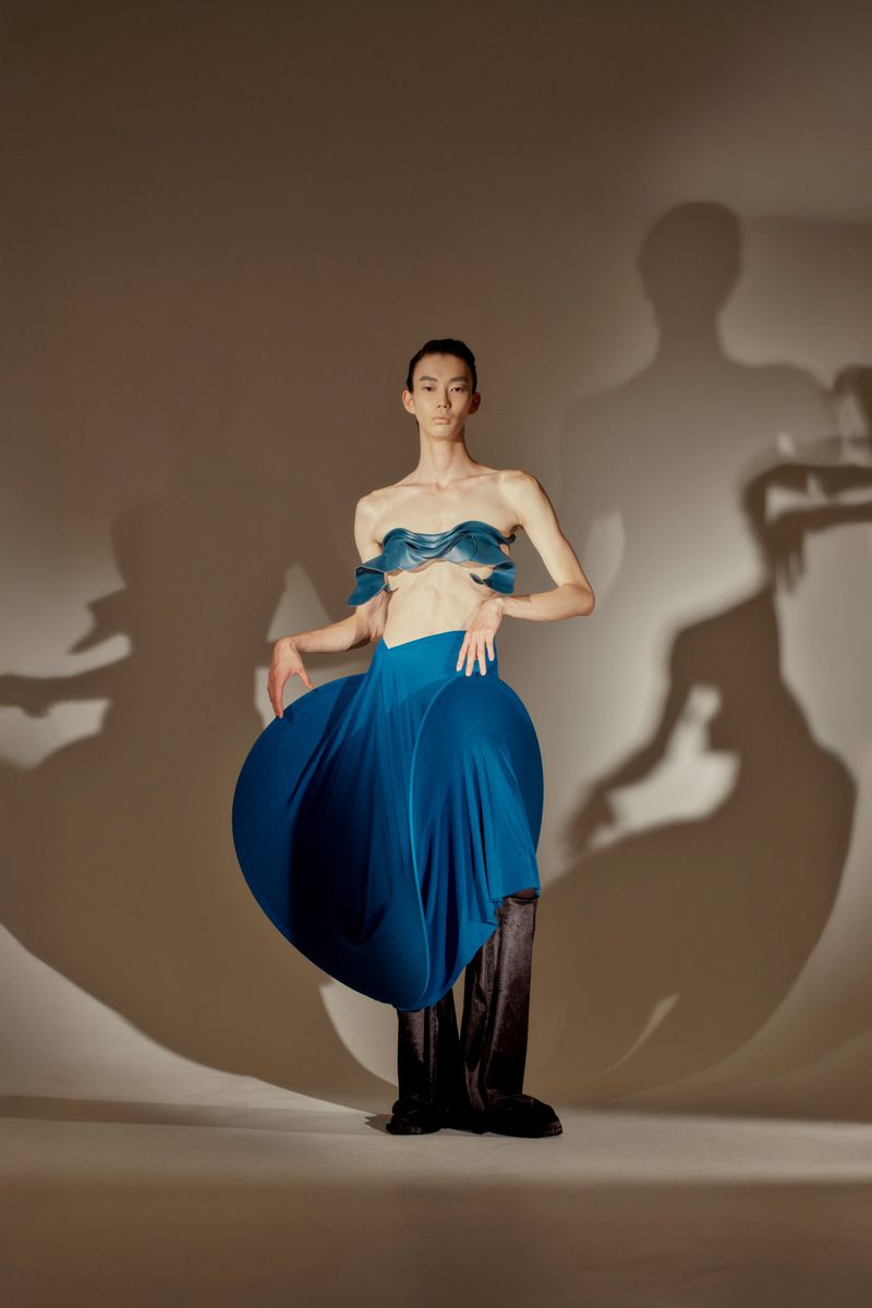 An Asian woman poses in a blue sculptural dress with dramatic shadows, shot in a studio with a neutral backdrop.