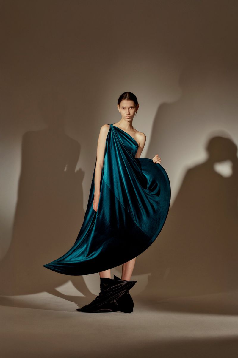 A slender woman poses in a flowing teal gown with asymmetrical neckline and black boots against a neutral background with soft shadows.