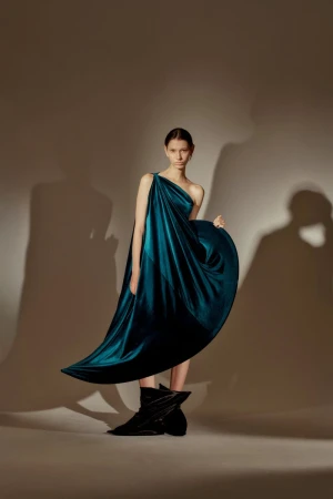A slender woman poses in a flowing teal gown with asymmetrical neckline and black boots against a neutral background with soft shadows.