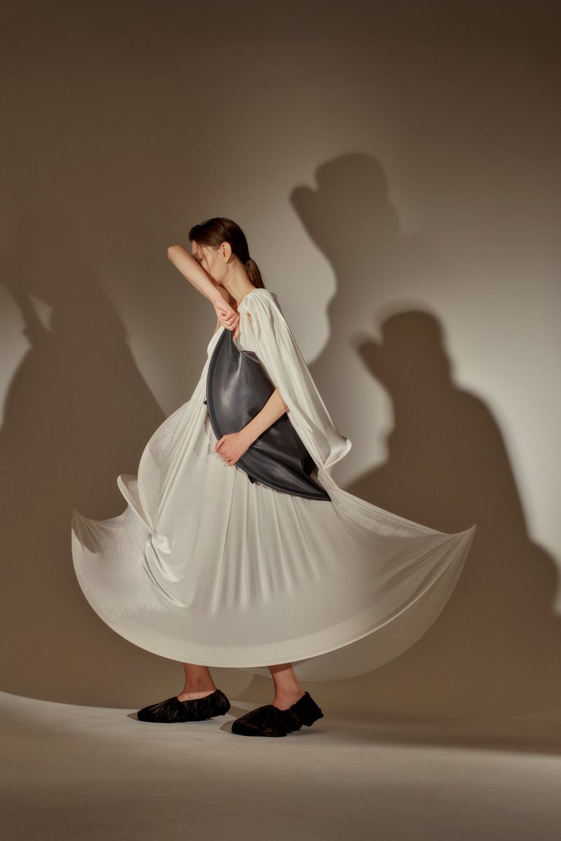A woman wearing a flowing white dress and black flats holds a black leather bag while posing in a neutral indoor setting with a shadow.