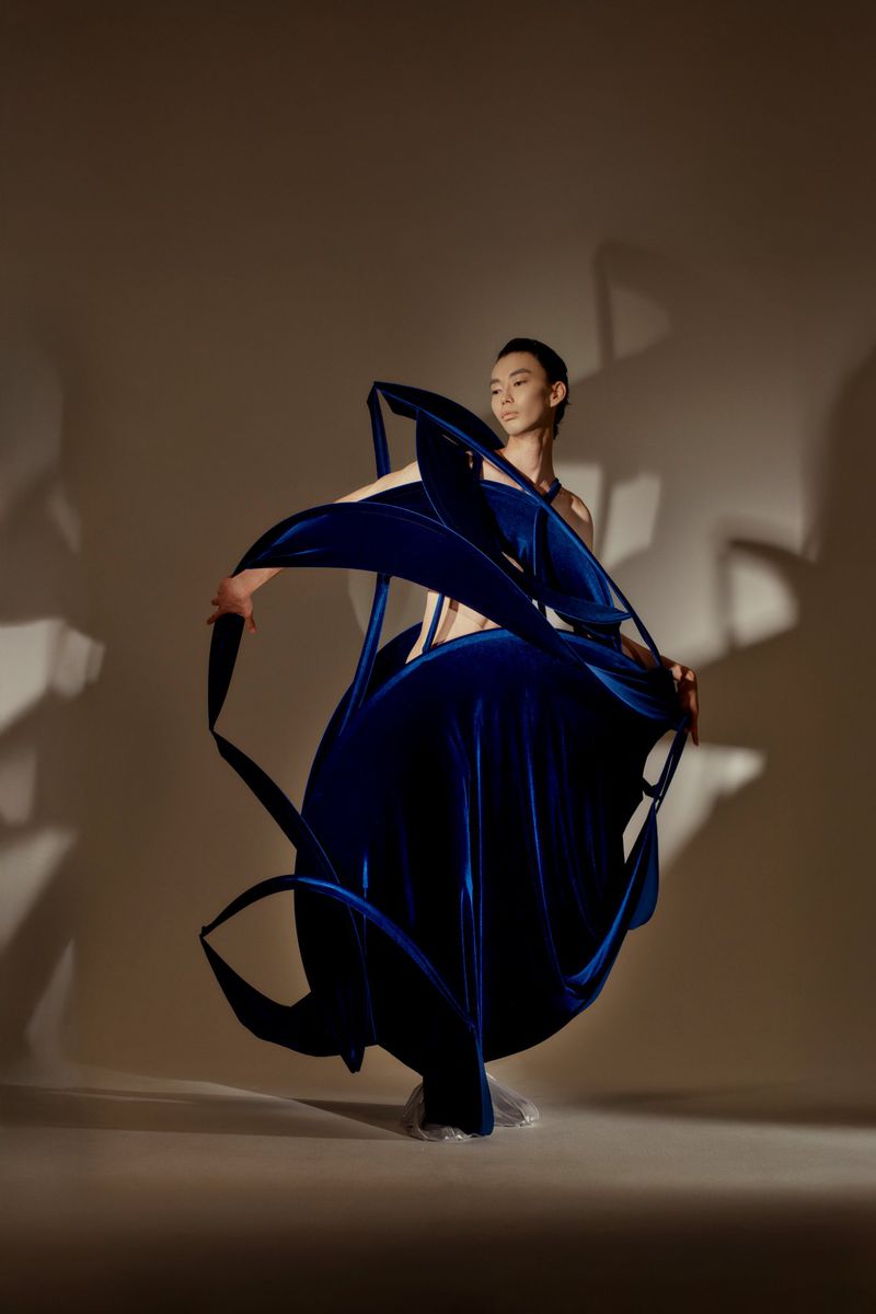 Asian woman wearing a large, sculptural, dark-blue velvet dress with flowing shapes, posing in a photo studio against a beige backdrop.