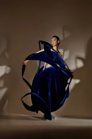 Asian woman wearing a large, sculptural, dark-blue velvet dress with flowing shapes, posing in a photo studio against a beige backdrop.