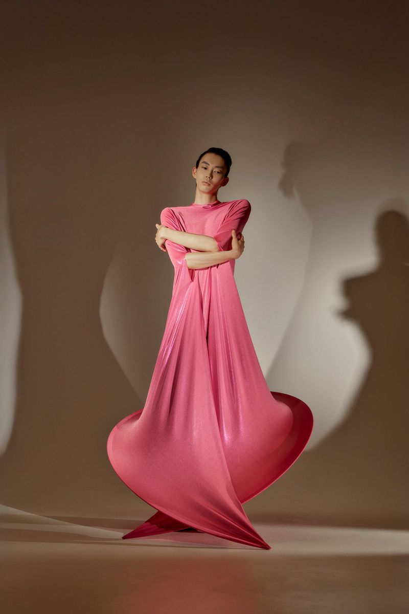Elegant woman in a flowing pink dress poses in a studio. The fabric creates dynamic shapes with dramatic lighting and shadows.