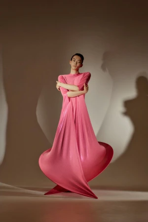 Elegant woman in a flowing pink dress poses in a studio. The fabric creates dynamic shapes with dramatic lighting and shadows.