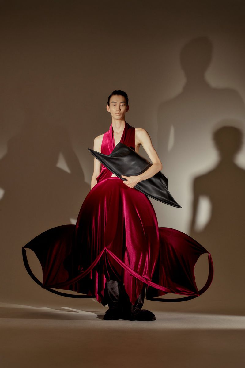 Fashionable person poses in red velvet dress with an angular black bag, standing against a neutral backdrop with projected shadows.