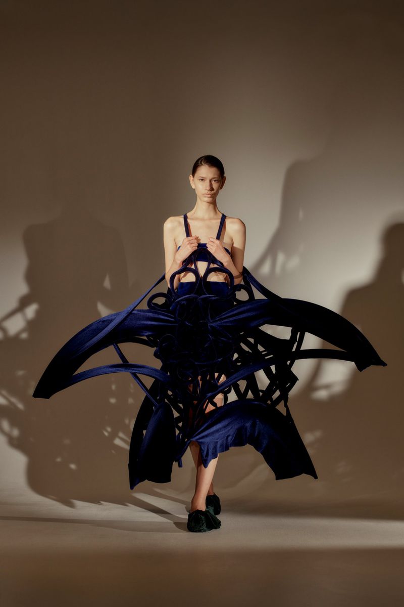 Fashion model in a blue, sculptural dress posing in a studio. The dress is an abstract design, and the model is facing the camera.