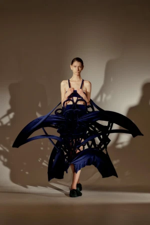 Fashion model in a blue, sculptural dress posing in a studio. The dress is an abstract design, and the model is facing the camera.