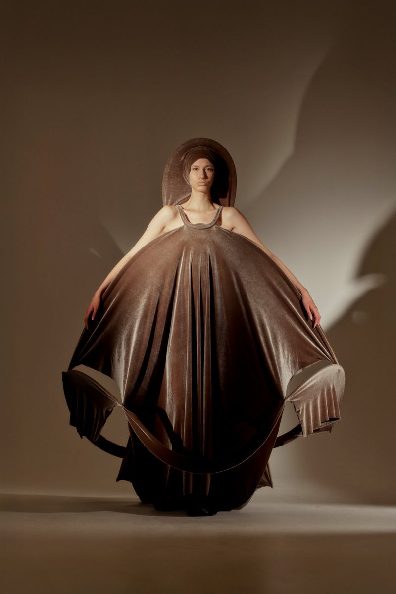 Woman in an artful, oversized tan dress with matching head covering poses in a photo studio with soft lighting and neutral background.