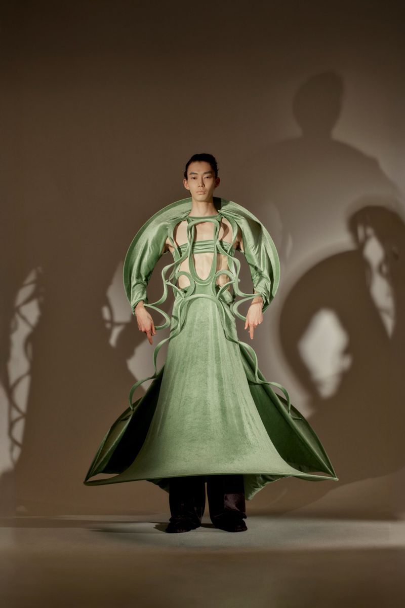 Fashion model showcases avant-garde green dress with structural design elements in a studio setting with a soft, neutral backdrop.
