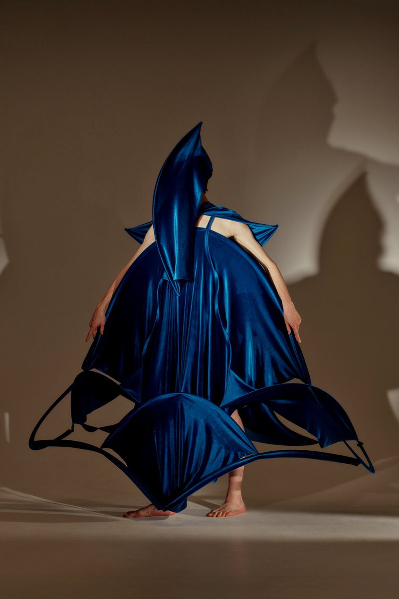A person in an elaborate, dark blue sculptural dress stands in a studio with a neutral background, creating an avant-garde fashion statement.