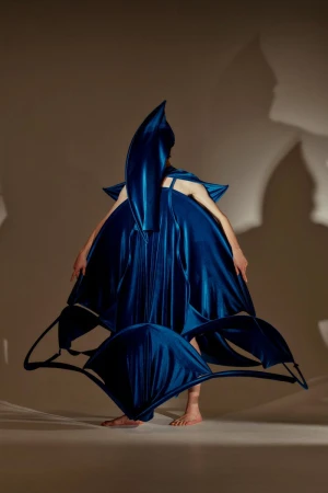 A person in an elaborate, dark blue sculptural dress stands in a studio with a neutral background, creating an avant-garde fashion statement.