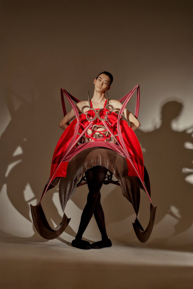 Fashion model showcases a dramatic red dress with unusual construction and large geometric shapes in a studio with cast shadows.