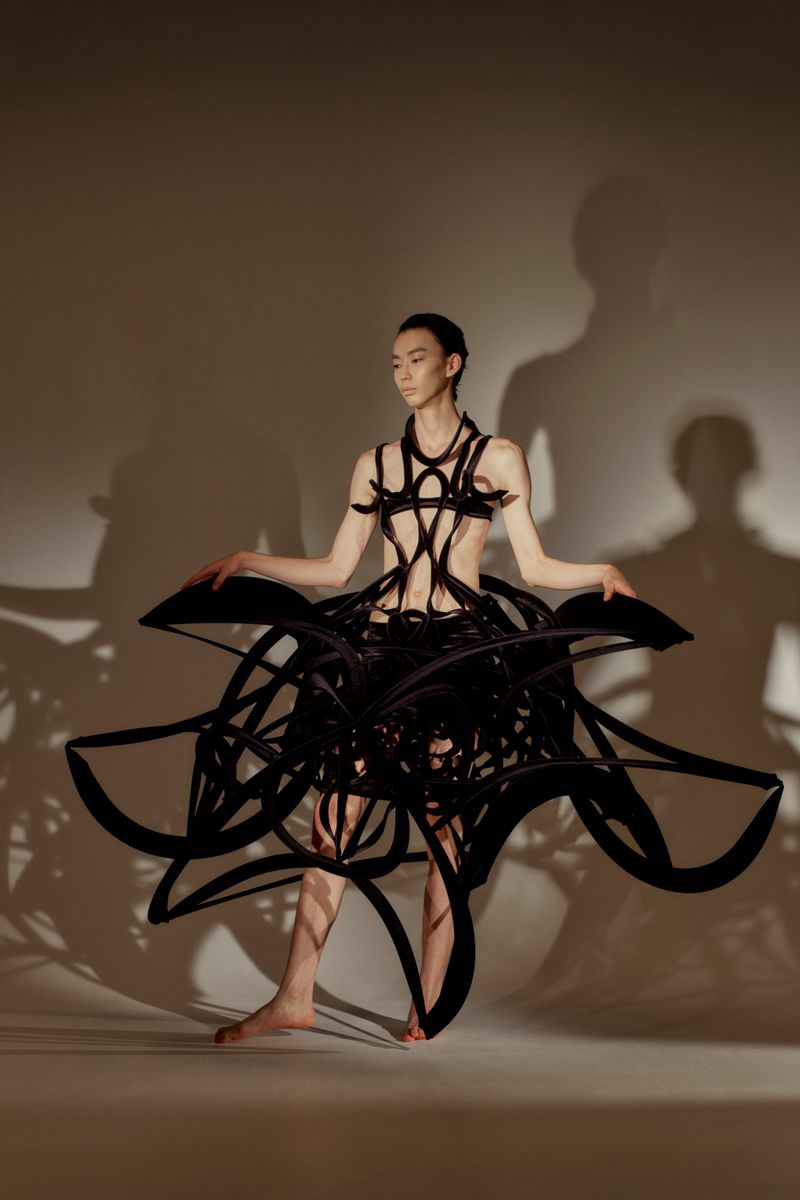 Asian woman poses in a striking black avant-garde fashion dress with abstract details, set against a neutral studio backdrop.