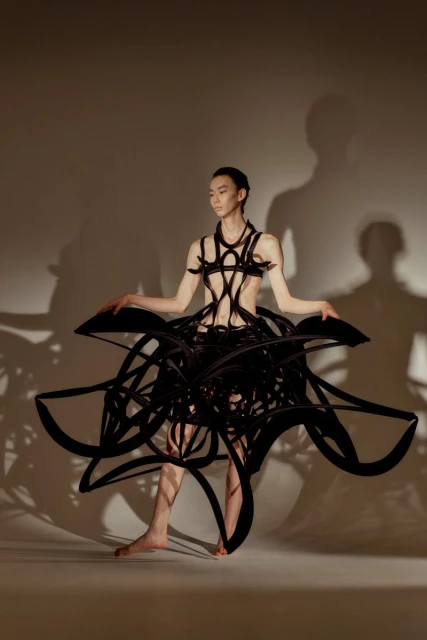 Asian woman poses in a striking black avant-garde fashion dress with abstract details, set against a neutral studio backdrop.
