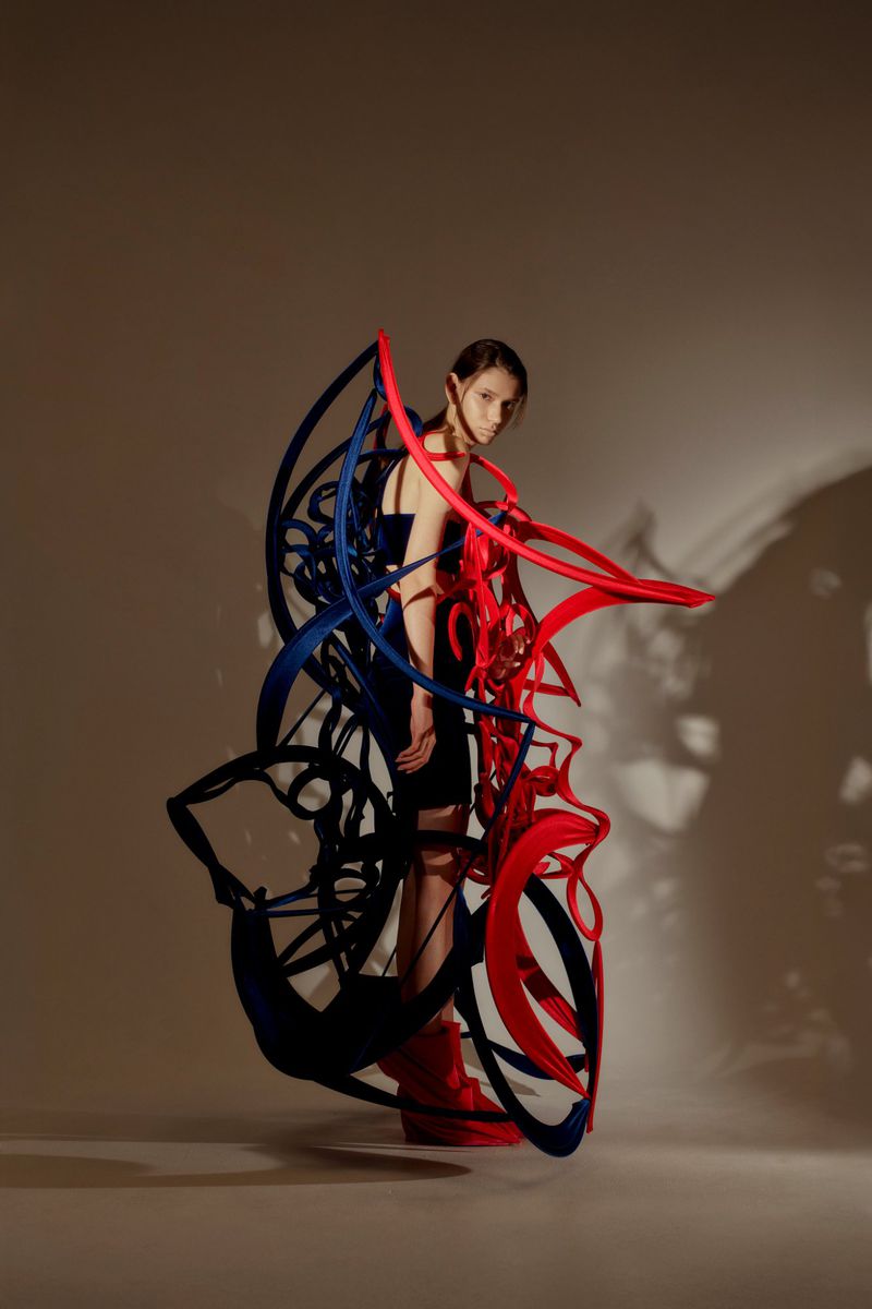 A fashion shot featuring a woman in a dark dress, partially obscured by a large sculptural garment of curved red and blue elements in a studio.